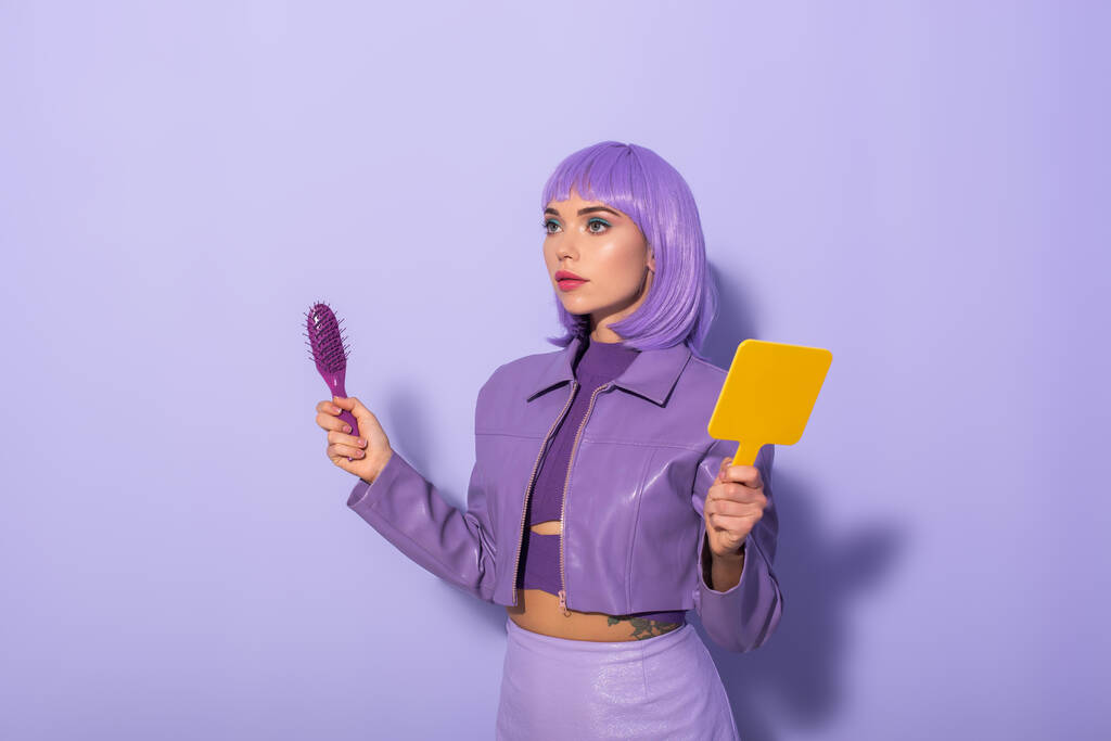 junge Frau im Puppenstil mit Haarbürste und Spiegel auf violettem Hintergrund gekleidet - Foto, Bild