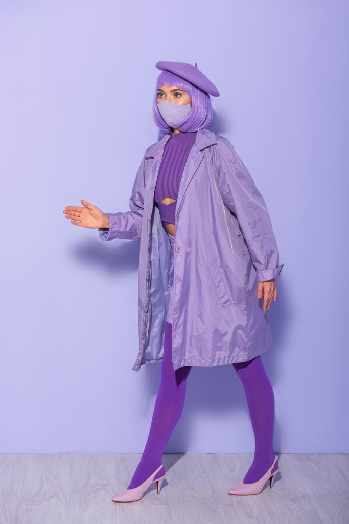 Nukketyyliin pukeutunut nuori nainen lääketieteellisessä naamiossa ja baskeri violetilla värikkäällä taustalla - Valokuva, kuva