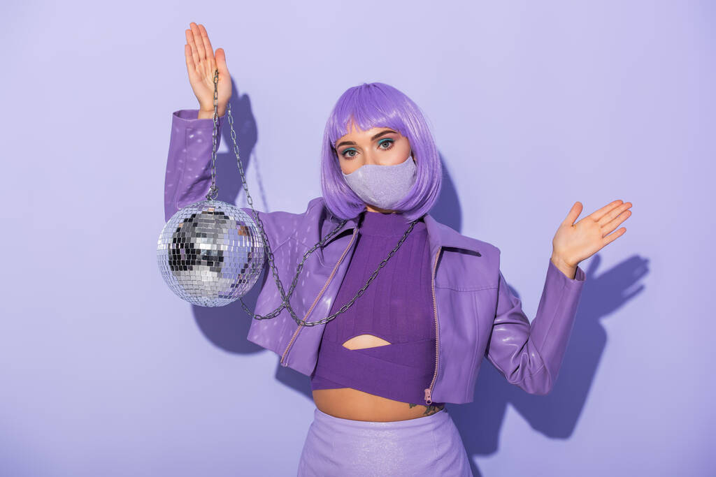 mujer joven vestida con estilo de muñeca en máscara médica con bola disco sobre fondo de color violeta - Foto, Imagen