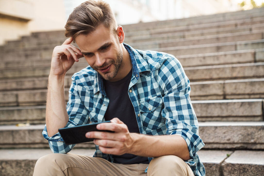 Handsome smiling young man using mobile phone while sitting on steps outdoors - Photo, Image