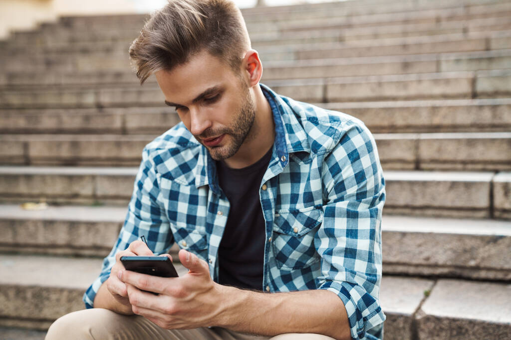 Guapo joven usando el teléfono móvil mientras está sentado en los escalones al aire libre - Foto, Imagen