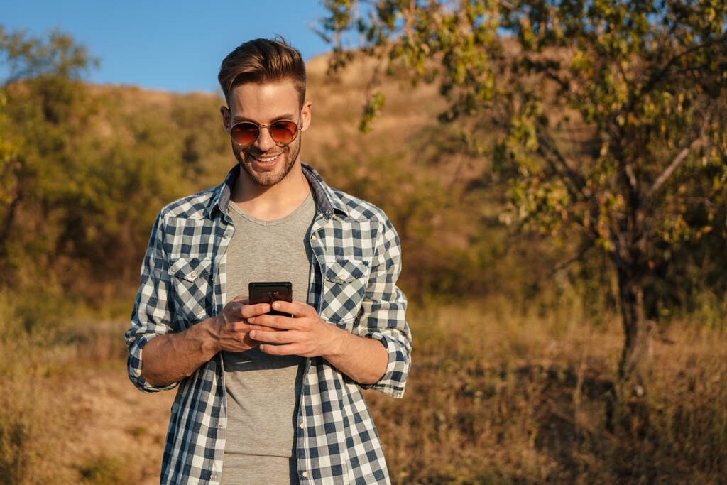 Bello ragazzo felice sorridente e utilizzando il cellulare mentre passeggiava sulla natura in estate - Foto, immagini