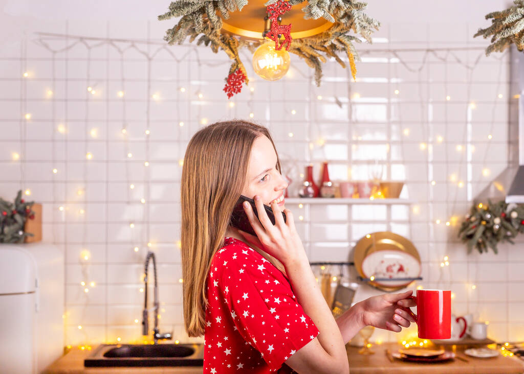 Щасливий сміх і усміхнена молода красива жінка говорить або розмовляє по мобільному телефону на кухні в червоних пігамах, тримаючи чашку чаю. Новий рік і Різдво вдома
. - Фото, зображення