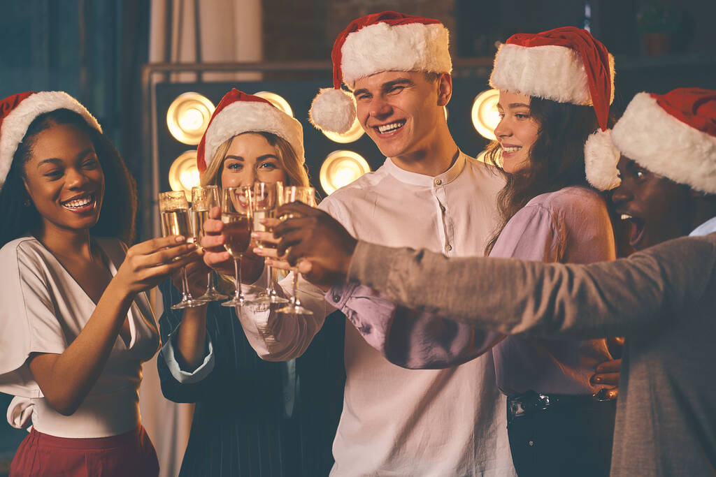 Hymyileviä miehiä ja naisia joulupukin hatuissa seisomassa samppanjalasien kanssa ja paahtamassa paahtoleipää. Hyvää uutta vuotta käsite - Valokuva, kuva