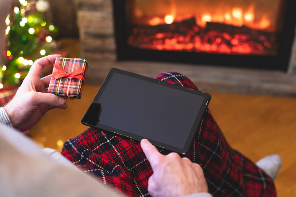 Navidad. Hombre usando la tableta para buscar ideas de regalo sentado con caja de regalo listo cerca de la chimenea y el árbol de Navidad. Concepto - Foto, Imagen