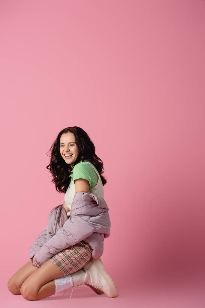 side view of happy brunette young woman in stylish winter outfit posing on knees on pink background - Photo, Image