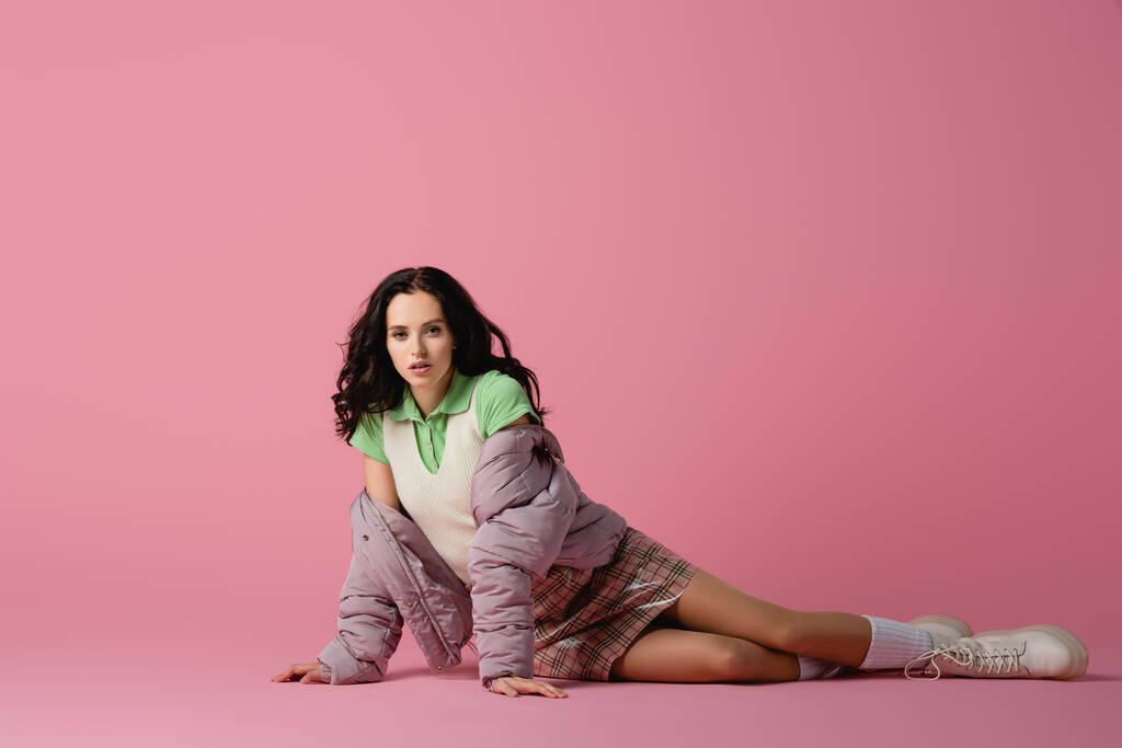 brunette young woman in stylish winter outfit posing on floor on pink background - Photo, Image