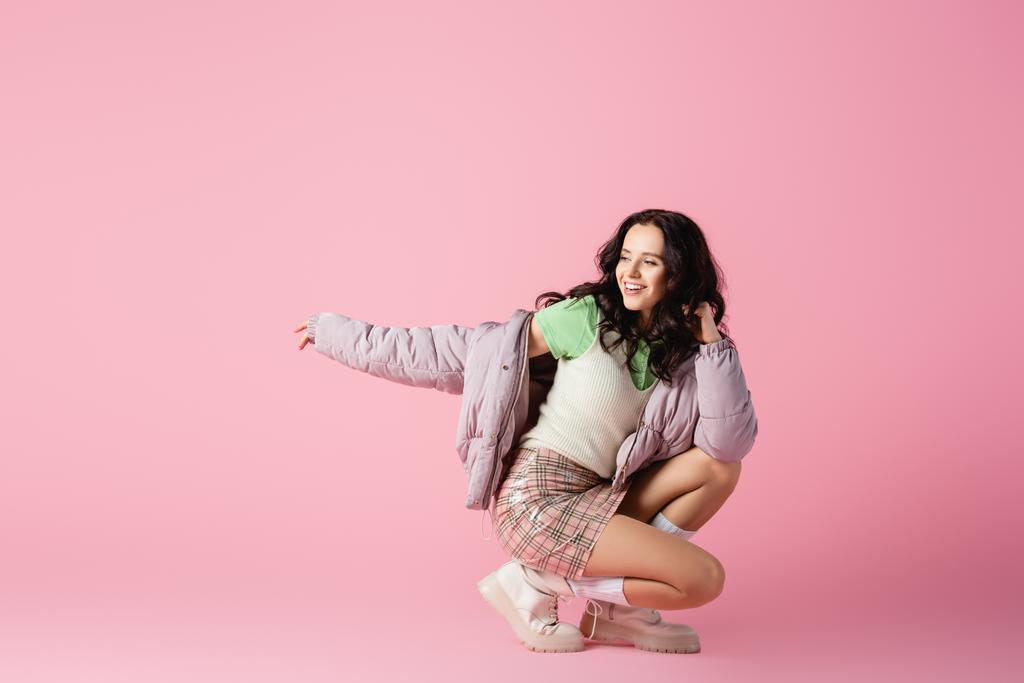 happy brunette young woman in stylish winter outfit posing on floor on pink background - Photo, Image