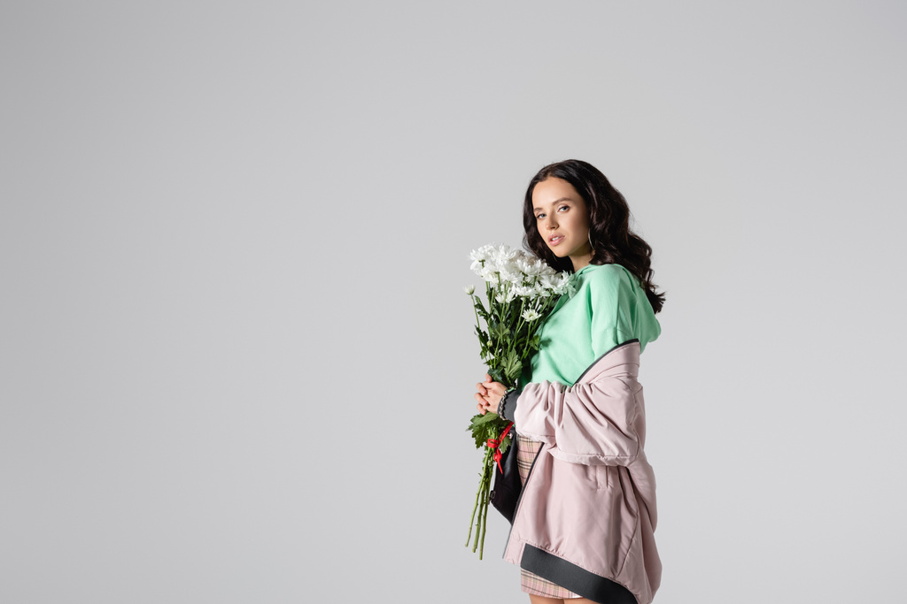 side view of brunette young woman in stylish winter outfit posing with flowers on grey background - Photo, Image