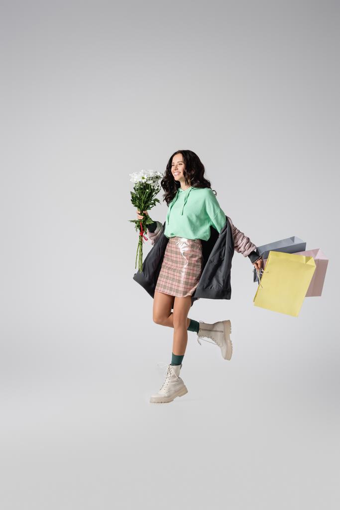 улыбающаяся брюнетка молодая женщина в стильном зимнем наряде позирует с цветами и сумками для покупок на сером фоне - Фото, изображение