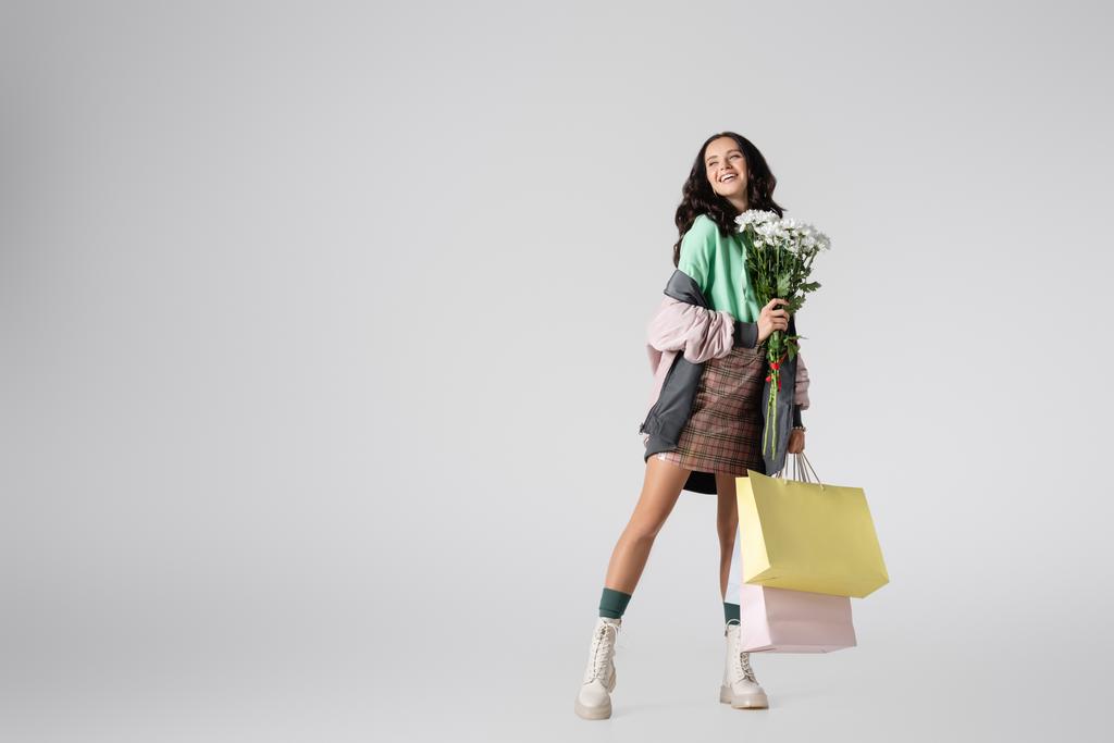 sonriente morena joven en elegante traje de invierno posando con flores y bolsas de compras sobre fondo gris - Foto, imagen
