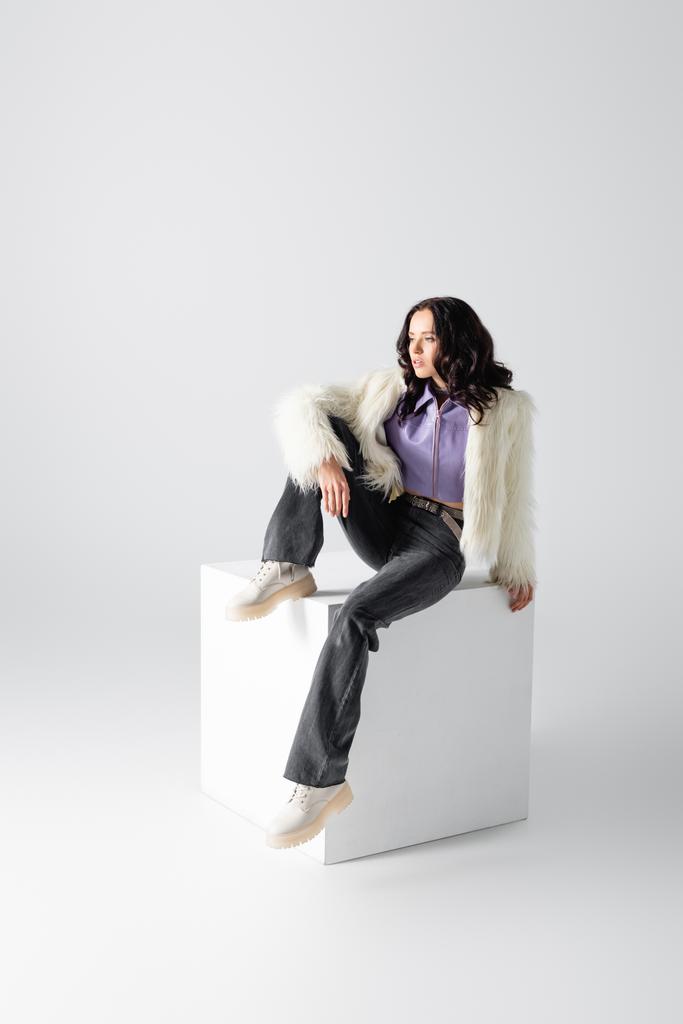 елегантна брюнетка молода жінка в стильній хутряній куртці позує на кубику на білому тлі
 - Фото, зображення