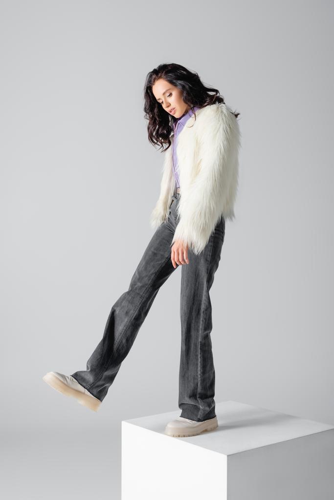 elegancki brunetka młoda kobieta w stylowy faux futro kurtka pozowanie na sześcian na białym tle - Zdjęcie, obraz