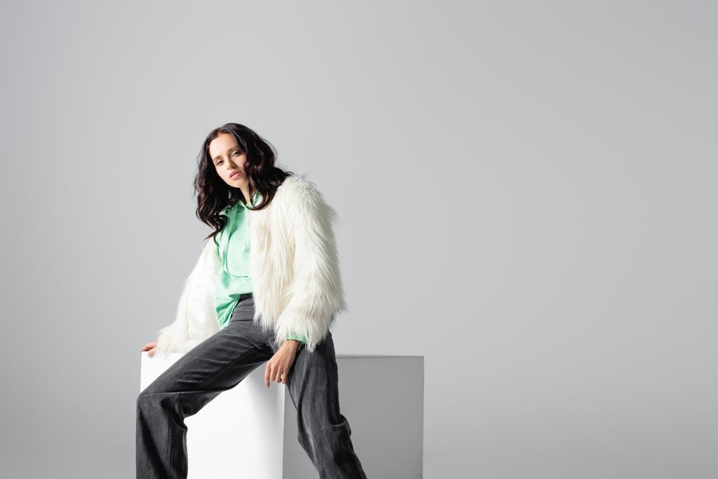 brunette jonge vrouw in faux fur jas poseren op kubus op witte achtergrond - Foto, afbeelding