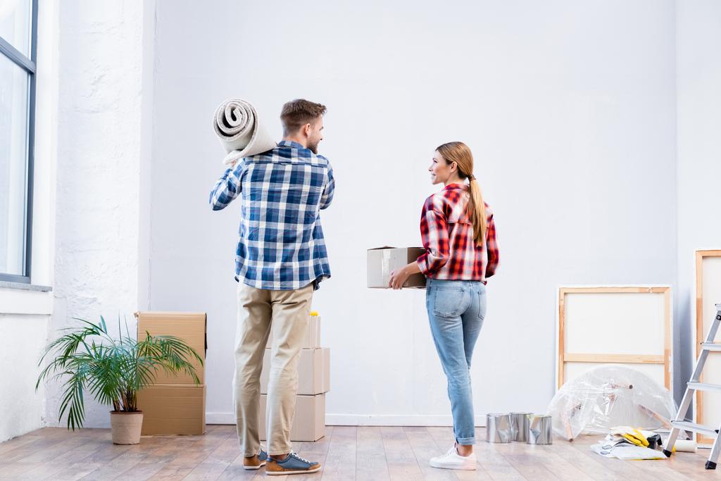 vista trasera de pareja joven sosteniendo caja de cartón y rollo de alfombra en casa - Foto, imagen