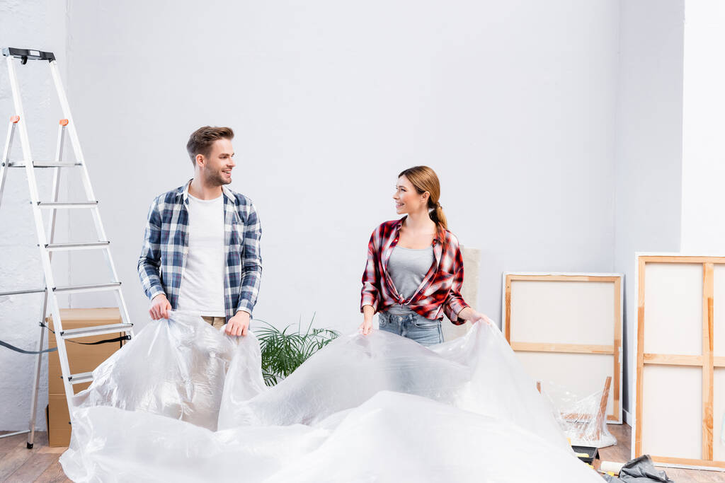 glückliches junges Paar mit Polyethylen, das sich anschaut, während es Zimmer während der Renovierung der Wohnung abdeckt  - Foto, Bild