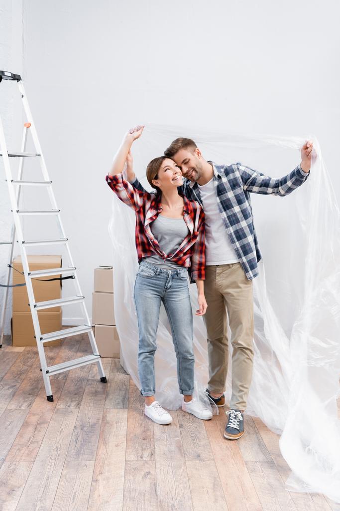 volle Länge des glücklichen jungen Paares, das sich umarmt, während es sich mit Polyethylen nahe der Leiter zu Hause bedeckt - Foto, Bild