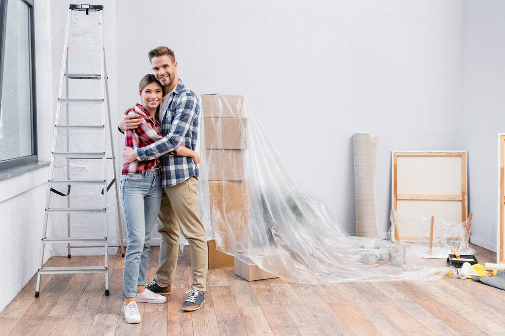 полная длина счастливой молодой пары смотрит в камеру, обнимаясь во время ремонта дома - Фото, изображение