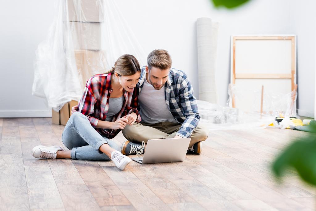 glückliches junges Paar blickt auf Laptop, während es zu Hause auf dem Boden auf verschwommenem Vordergrund sitzt - Foto, Bild