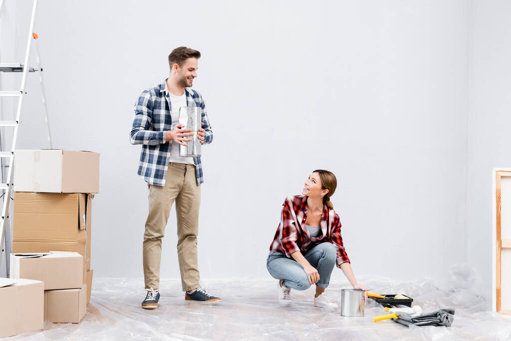 comprimento total do jovem sorridente com espátula olhando para a mulher sentada perto de rolo de pintura no chão dentro de casa - Foto, Imagem