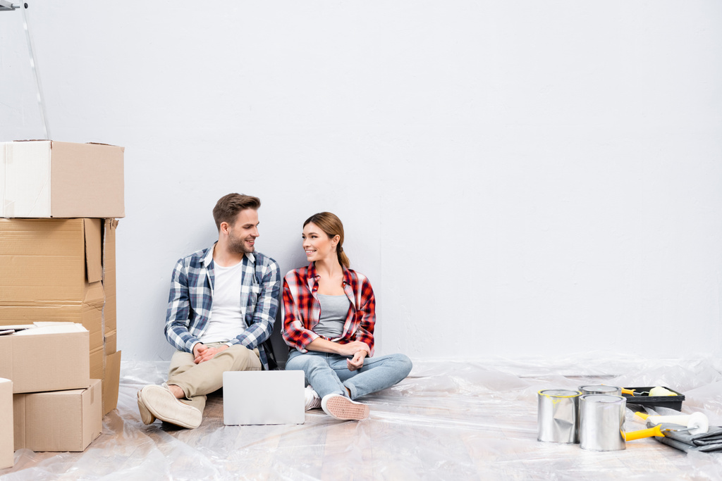 full length of happy young couple looking at each other while sitting near laptop and cardboard boxes on floor at home - Photo, Image