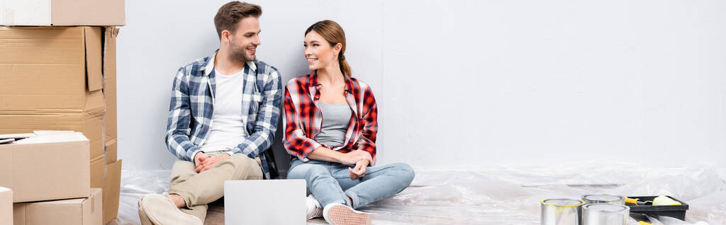 улыбающаяся молодая пара смотрит друг на друга, сидя рядом с ноутбуком и картонными коробками на полу дома, баннер - Фото, изображение