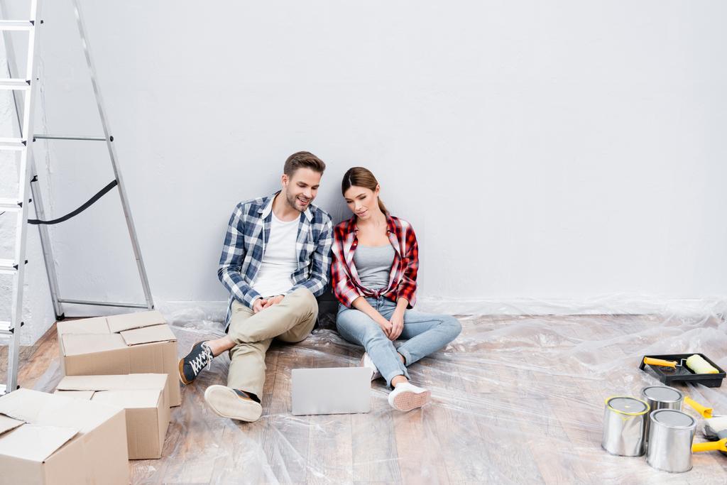 full length of happy young couple looking at laptop while sitting on floor near cardboard boxes, blikken verf en ladder thuis - Foto, afbeelding