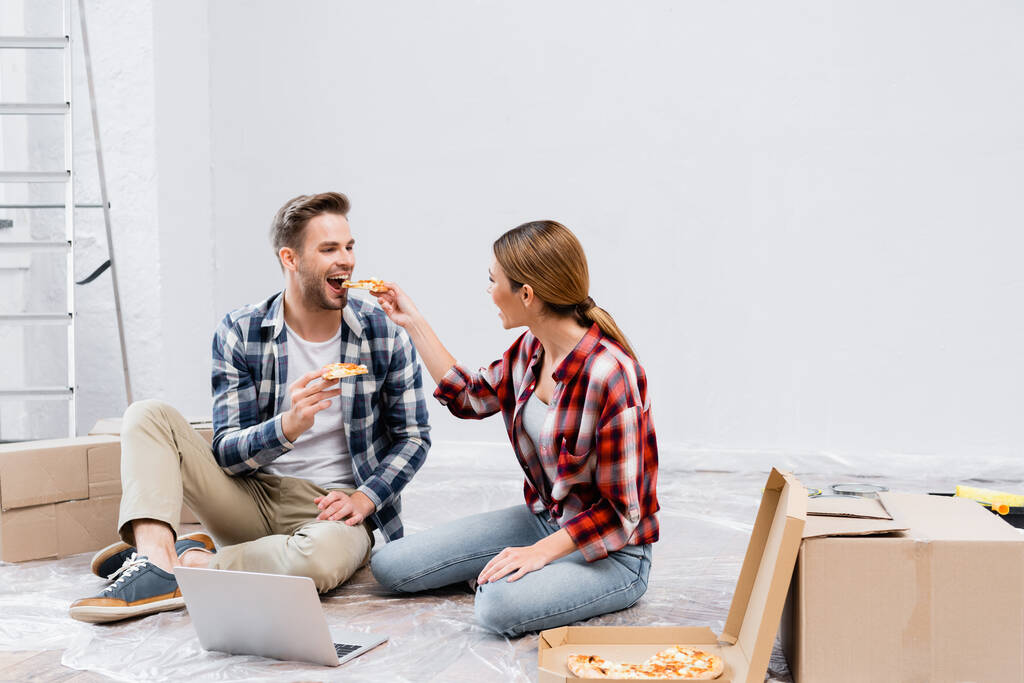 自宅のラップトップの近くの床に座っている間に幸せな女性がピザを食べる男の完全な長さ - 写真・画像