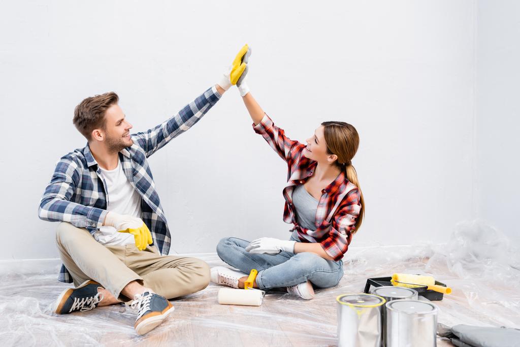 幸せな若いカップルの完全な長さ手袋を与え、互いに高い5を与えながら床の上に座っている家で塗料の缶の近くに - 写真・画像