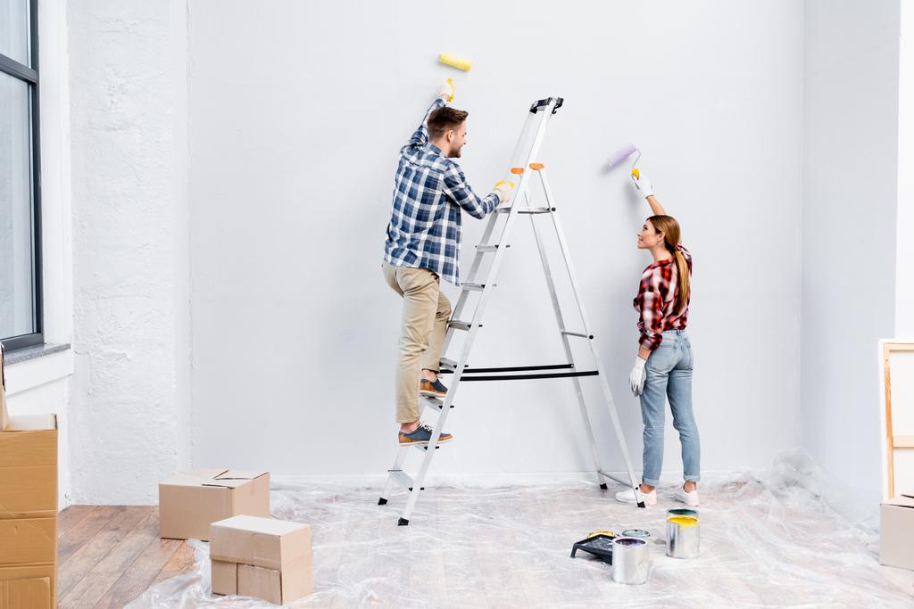 comprimento total de casal jovem sorridente com rolos de parede de pintura e olhando uns para os outros perto escada em casa - Foto, Imagem