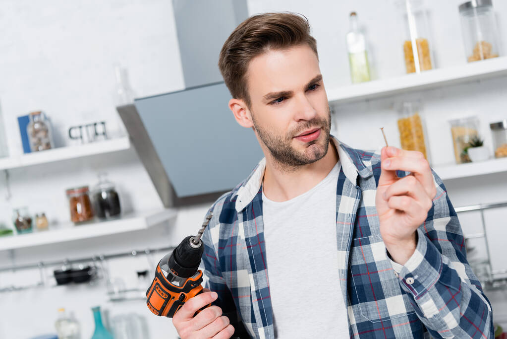 young man with drill looking at nail on blurred background in kitchen - Photo, Image