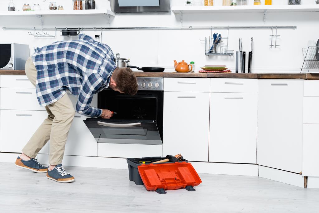 pełna długość młodego człowieka ze szczypcami zginającymi się w pobliżu piekarnika i skrzynki narzędziowej w kuchni - Zdjęcie, obraz
