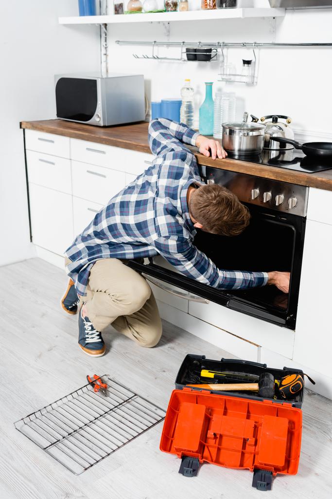 volledige lengte van de jongeman repareren oven terwijl zitten op de vloer in de buurt van raster en gereedschapskist in de keuken - Foto, afbeelding
