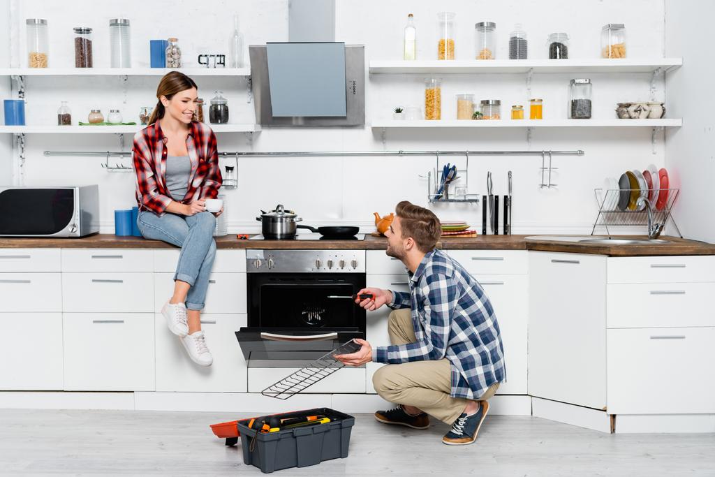 comprimento total da mulher sorridente com xícara de café olhando para o homem com grade perto do forno e kit de ferramentas na cozinha - Foto, Imagem