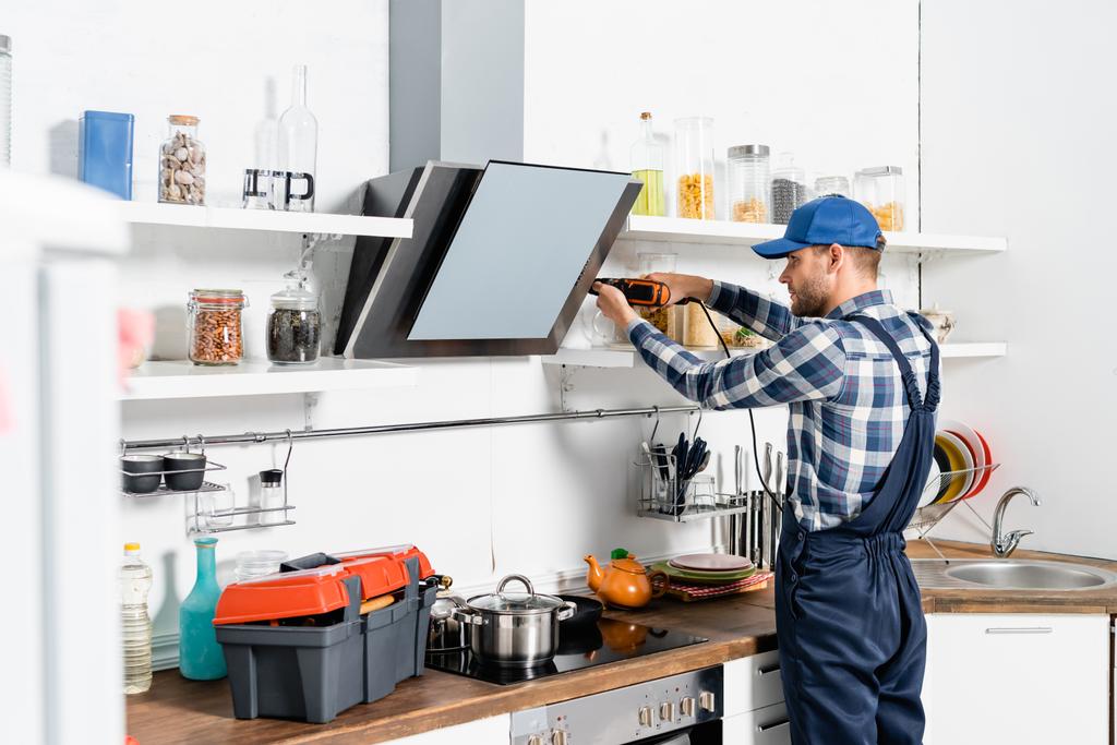 молодой мастер с помощью дрели рядом с вентилятором экстрактора на размытом переднем плане на кухне - Фото, изображение