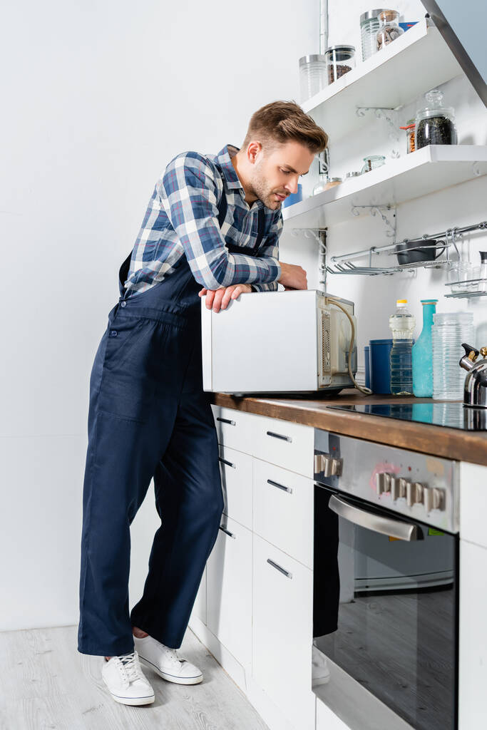 повна довжина молодого ремонтанта, який дивиться на мікрохвильову піч на кухні
 - Фото, зображення
