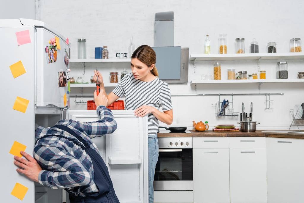νεαρή γυναίκα δίνει κατσαβίδι σε μάστορα επισκευή ψυγείου στην κουζίνα - Φωτογραφία, εικόνα