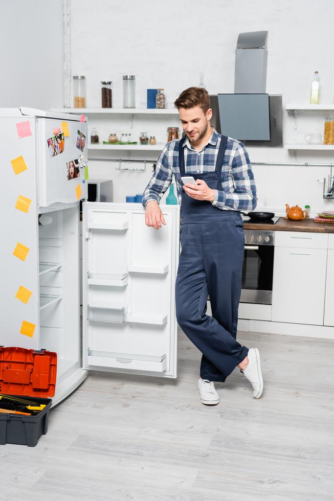 plná délka smajlíka údržbář smskování na smartphone zatímco se opírají o ledničku v kuchyni - Fotografie, Obrázek