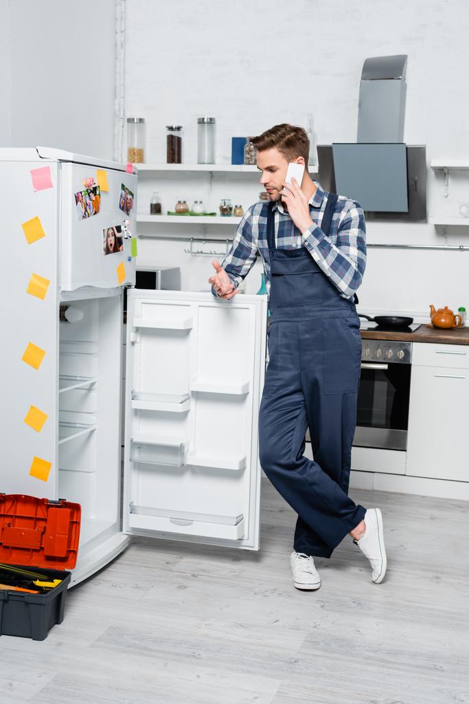 volledige lengte van de jonge reparateur praten op smartphone, terwijl leunen op de koelkast in de buurt gereedschapskist in de keuken - Foto, afbeelding