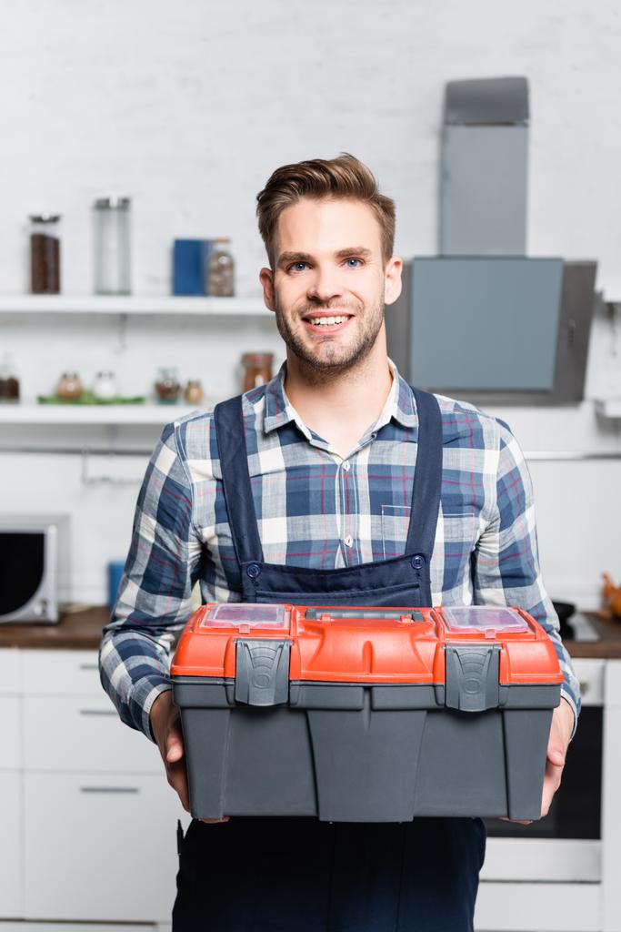 Vorderseite des glücklichen Reparateurs, der in die Kamera schaut, während er Werkzeugkoffer mit verschwommener Küche auf dem Hintergrund hält - Foto, Bild