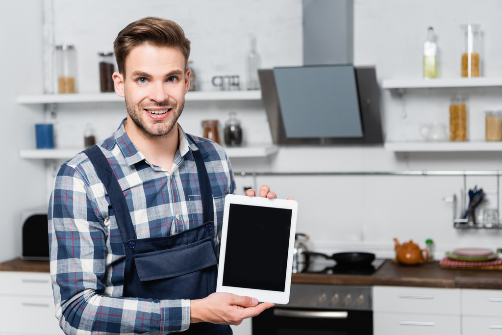 přední pohled šťastný údržbář při pohledu na kameru, zatímco ukazuje tablet s rozmazanou kuchyní na pozadí - Fotografie, Obrázek