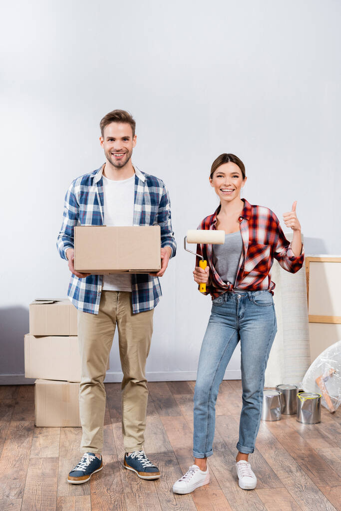полная длина счастливой молодой женщины с краски ролика показывая большой палец вверх рядом с мужчиной с картонной коробкой дома - Фото, изображение