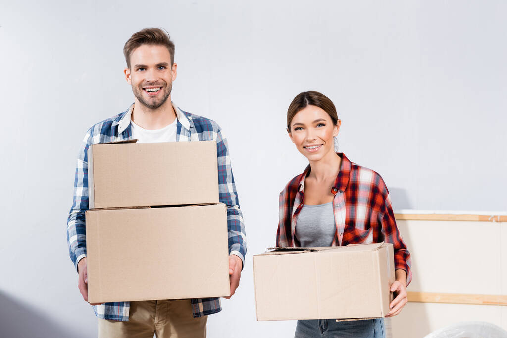 front view of cheerful young couple looking at camera while holding cardboard boxes at home - Photo, Image