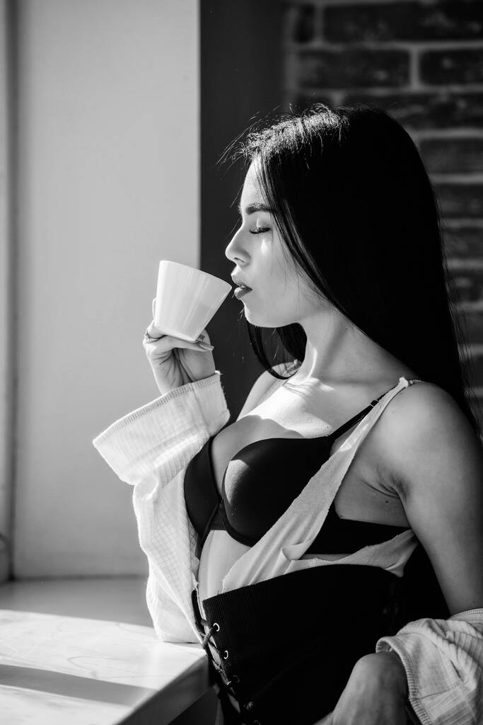 Genieten van ochtendzonlicht. Sensueel meisje sexy borsten drinken koffie in de buurt van vensterbank. Aantrekkelijke vrouwelijke lingerie ontspannen thuis. Gepassioneerde mysterieuze minnaar. Perfecte koffie tijd. Maar eerst koffie. - Foto, afbeelding