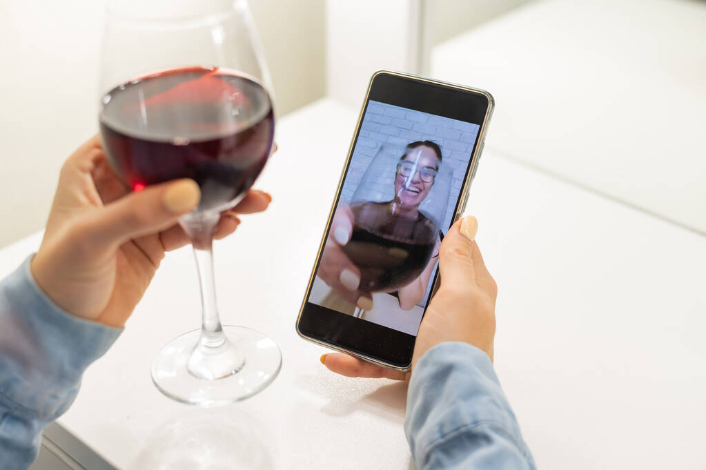 Οι γυναίκες μιλάνε μέσω βίντεο στο τηλέφωνο και πίνουν κόκκινο κρασί. Το κορίτσι σε καραντίνα δεν κάνει βιντεοκλήση. - Φωτογραφία, εικόνα