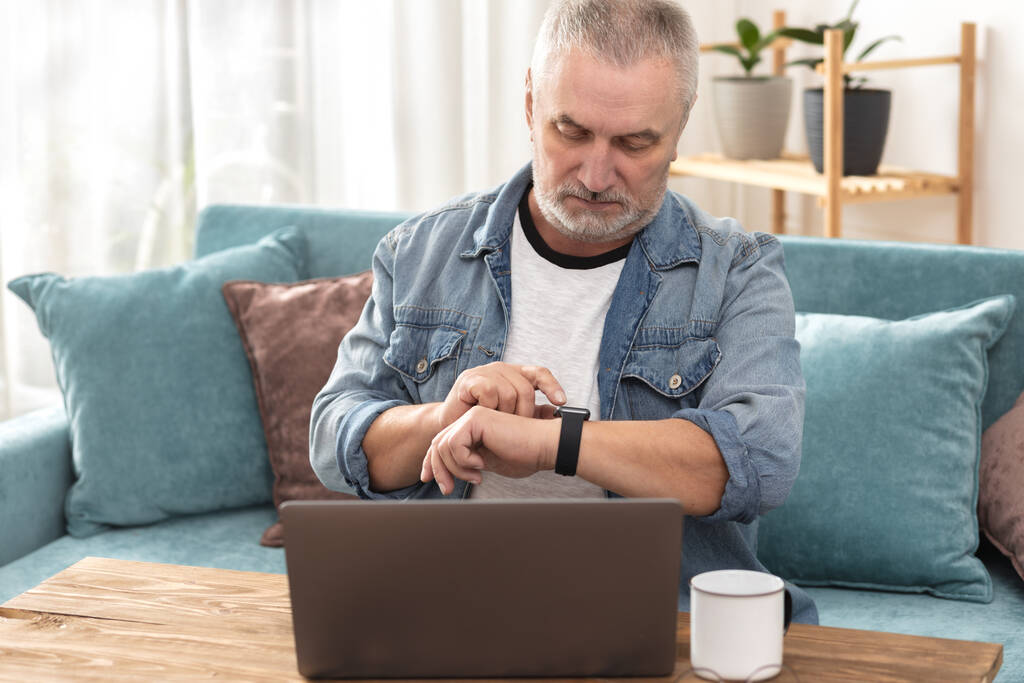 Uomo anziano si siede sul divano al suo computer portatile e riceve il messaggio sul suo orologio intelligente - Foto, immagini