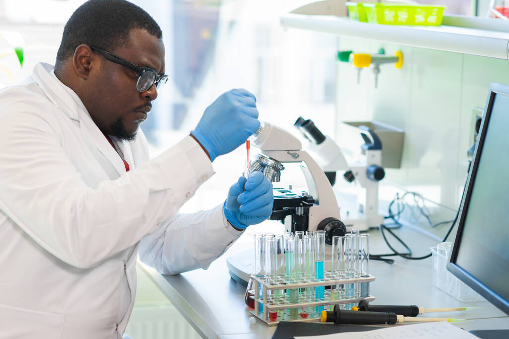 Афро-американский ученый работает в лаборатории. Доктор проводит микробиологические исследования. Лабораторные инструменты: микроскоп, пробирки, оборудование. Биотехнологии, химия, бактериология, вирусология, ДНК и здоровье - Фото, изображение