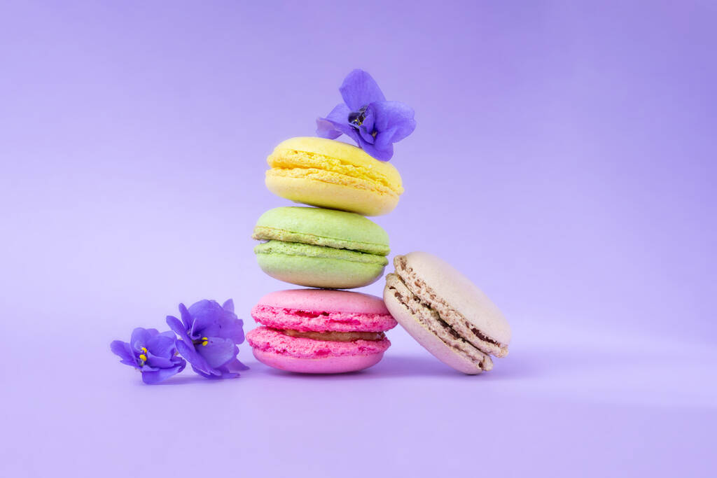 Macarrones coloridos con flores uno encima del otro sobre fondo púrpura. Galletas francesas pastelería tradicional - Foto, imagen