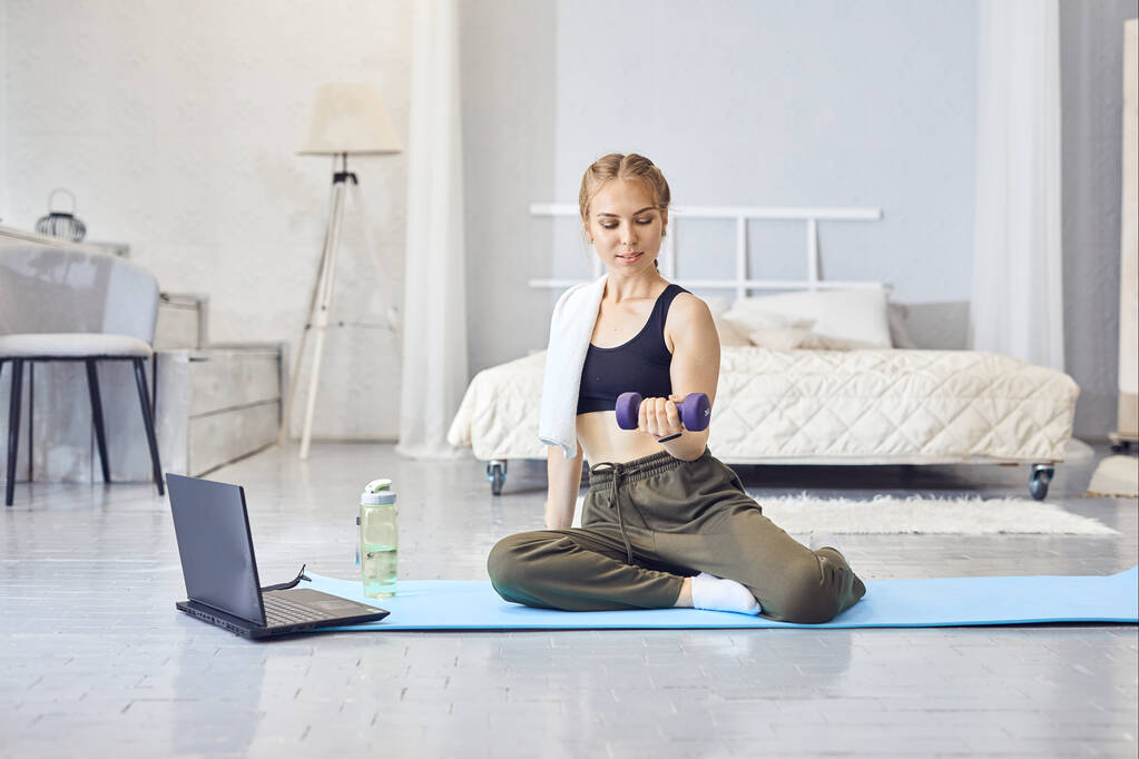 Eine sportliche Frau in Sportkleidung trainiert und nutzt zu Hause im Wohnzimmer einen Laptop. Fitnesskonzept. - Foto, Bild