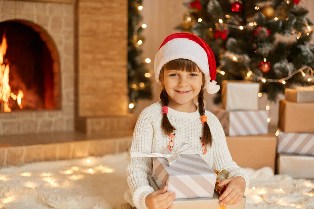 Piękna uśmiechnięta dziewczynka z prezentami siedząca na podłodze i patrząca w kamerę, nosząca biały sweter i czerwony kapelusz, dziecko pozujące w pobliżu kominka i choinki. - Zdjęcie, obraz